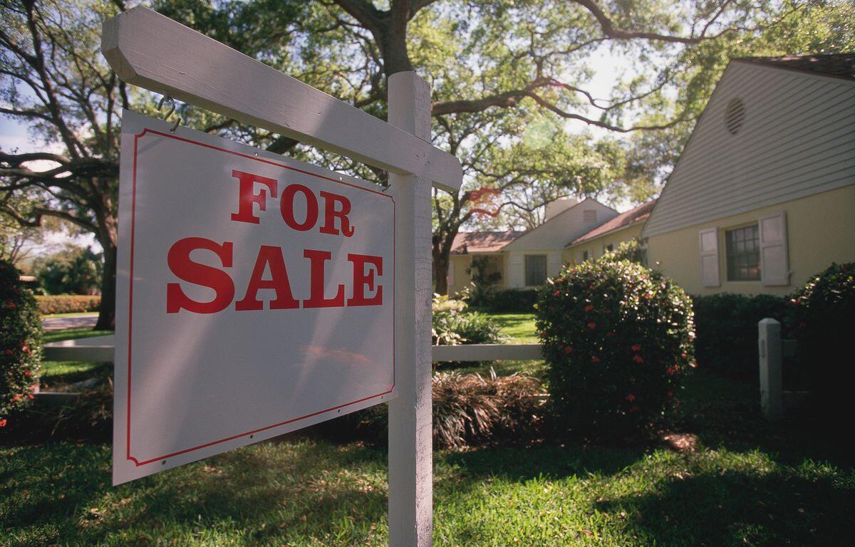 White wooden 'For Sale' sign in the front yard of a suburban house with trees behind it.
