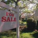 White wooden 'For Sale' sign in the front yard of a suburban house with trees behind it.