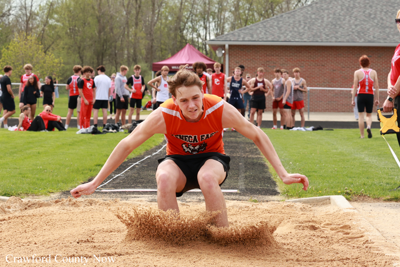 Male athlete in orange uniform lands in a sand pit during a track meet, with spectators watching in the background.