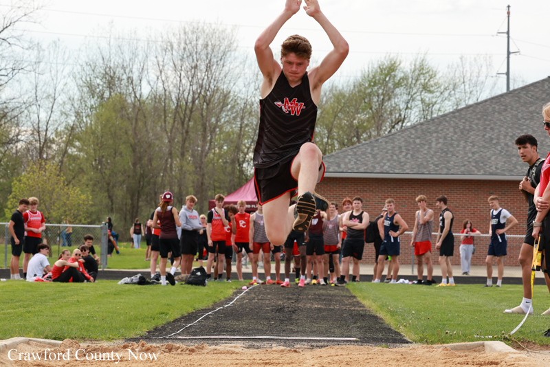Male athlete in a black singlet clears a high jump bar mid-air over the sand pit, with a crowd of teammates and spectators watching in the background.