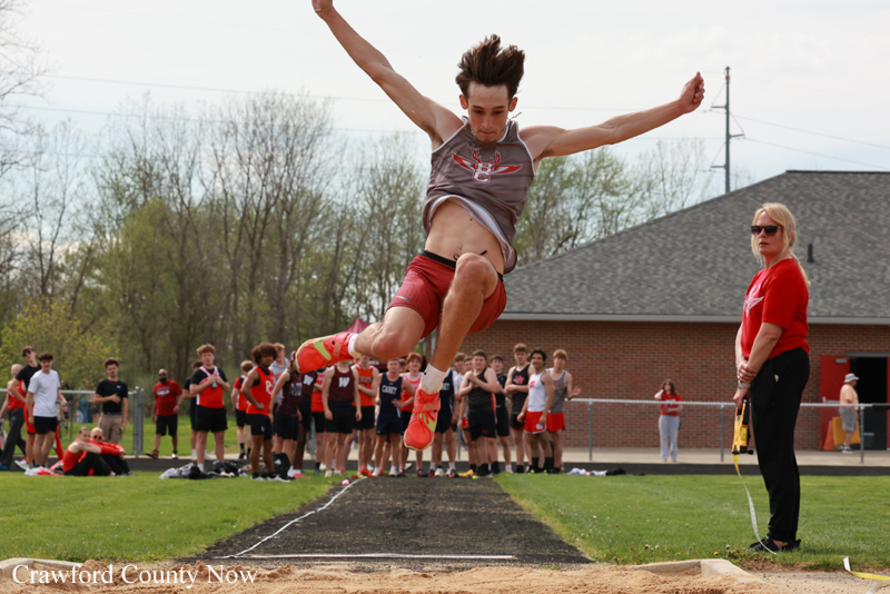 Male athlete jumps mid-air during a long jump at a track meet, with a crowd and official watching near the sand pit.