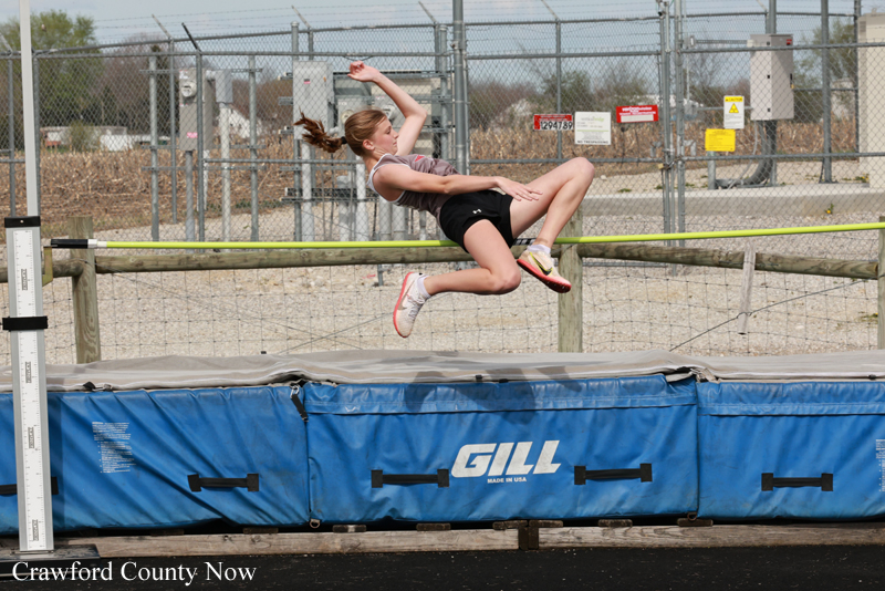 Female athlete midair performing a high jump over a padded blue mat outdoors, fence and equipment visible in the background.