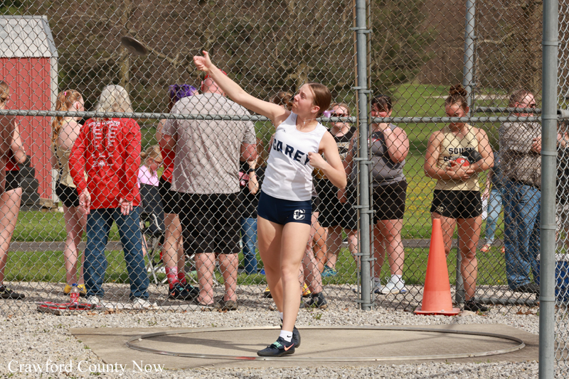 Female athlete in a white tank top and navy shorts releases a shot put inside a throwing circle as spectators watch behind a chain-link fence behind her.