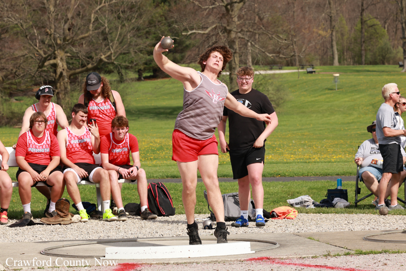 Male hammer thrower in gray tank top and red shorts releases the hammer from a throwing circle as teammates in red uniforms and onlookers watch in a park setting.