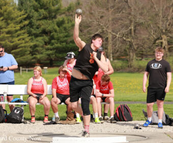Athlete in black shirt throws a shot put while a group watches at a grassy outdoor field event.