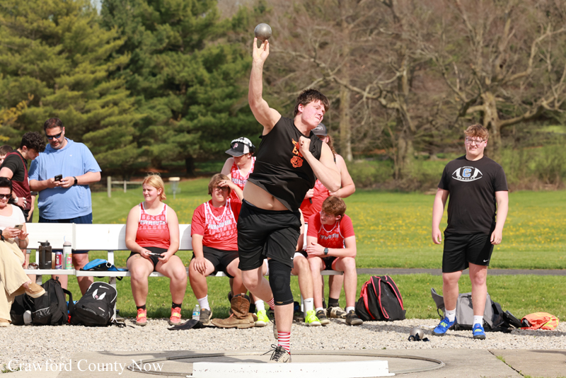 Athlete in black shirt throws a shot put while a group watches at a grassy outdoor field event.