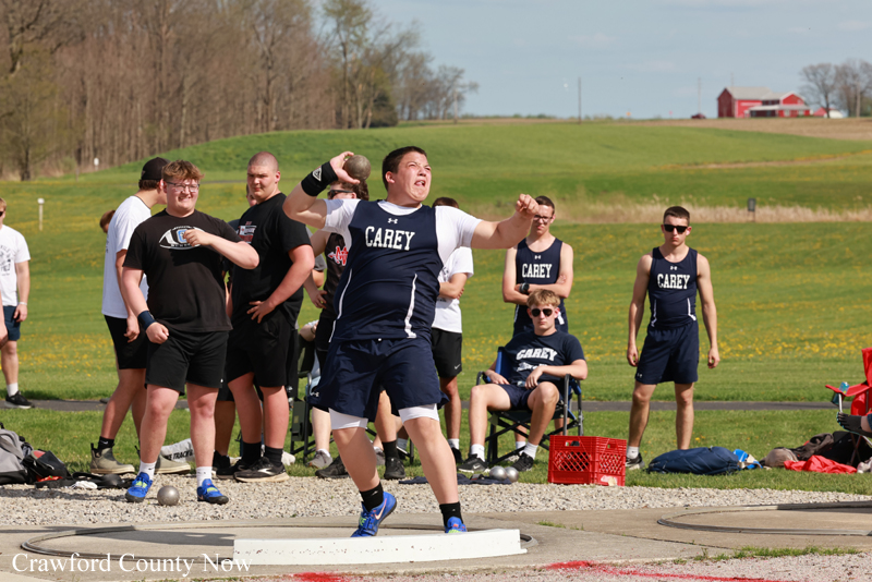 Male athlete in a navy Carey uniform mid-shot put throw on the throwing circle, teammates watching in a sunny field.
