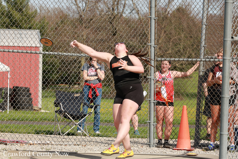 Female discus thrower in black athletic wear mid-throw near a chain-link fence, with spectators watching behind the fence.