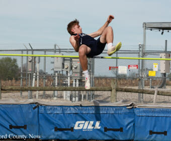 Male athlete mid-air clears a yellow high jump bar over a blue padded pit with 'GILL' branding in the foreground.