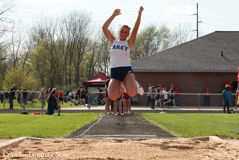 Female track athlete in a white and navy uniform is mid-air clearing the high jump bar during a meet, with a sand pit in the foreground and spectators behind a chain-link fence.