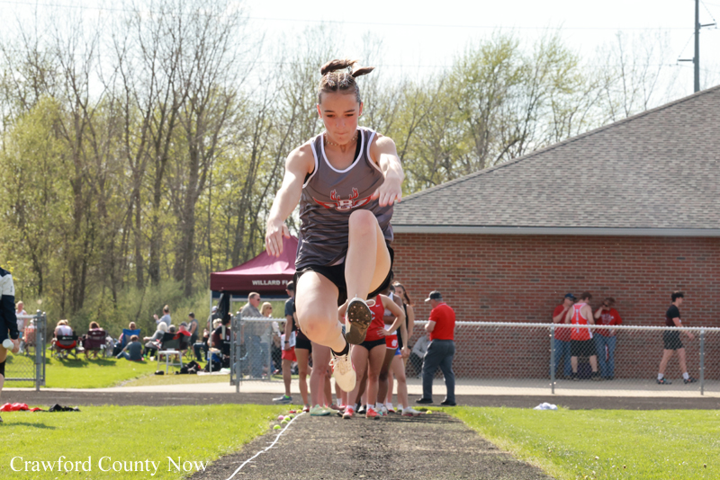 Female athlete midair performing a long jump on an outdoor track, with spectators and a red brick building in the background.