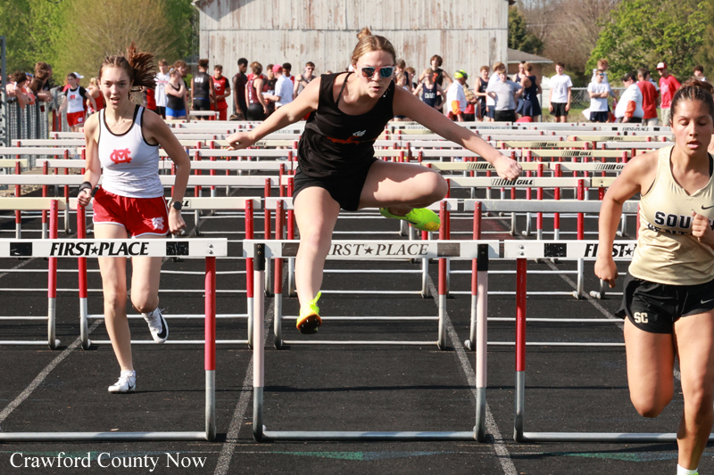 Female hurdlers mid-race clearing hurdles during a track meet; central runner in black jumps over a hurdle while two competitors follow.