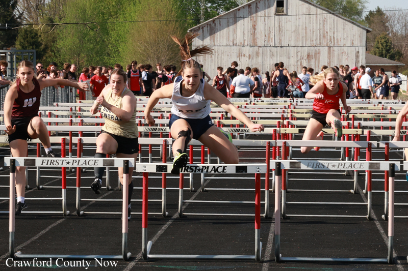 Four female hurdlers leap over hurdles in a crowded track meet, with a barn and spectators in the background.