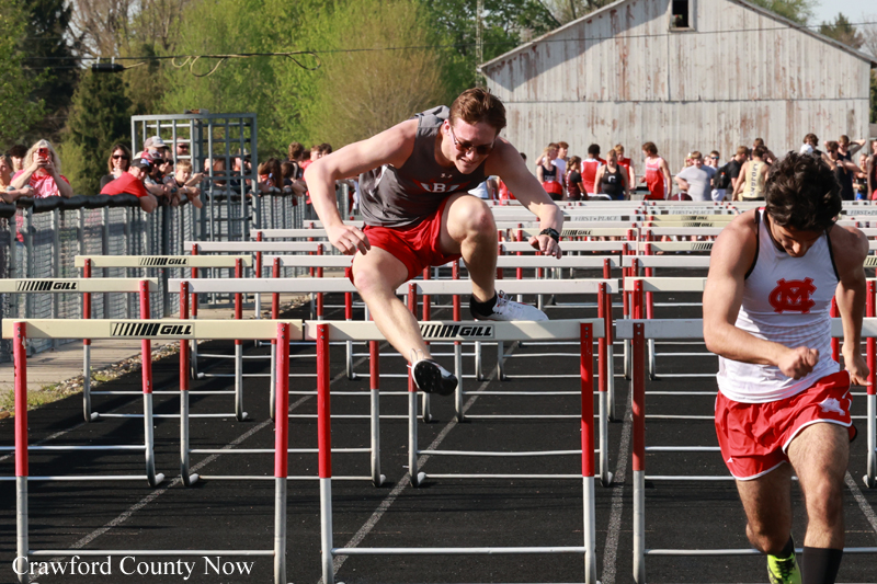 Male hurdler in gray top and red shorts leaps over a hurdle during a track race as spectators watch from behind the fence, on a sunny day by Crawford County Now audience area.