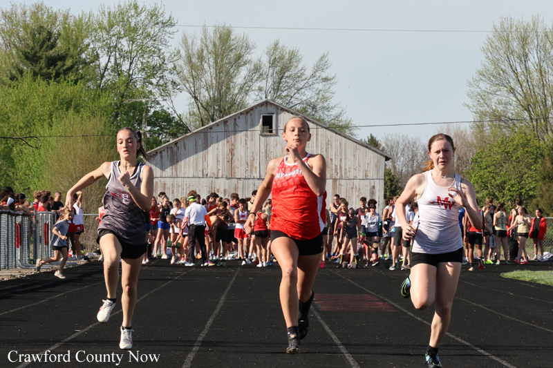Three female runners sprint from the start on a track, crowd cheering in the background near a barn, spring foliage around.