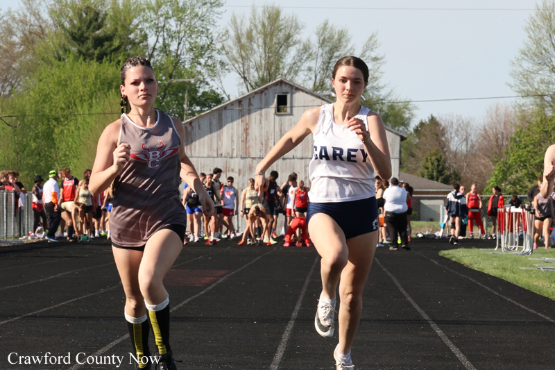 Two female sprinters race toward the camera on a track, with spectators and a barn-like building in the background.