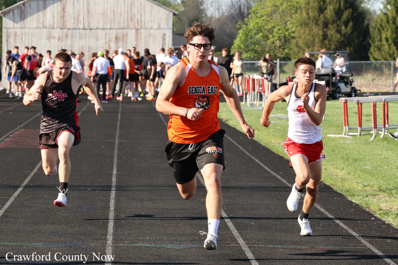 Three male sprinters race on an outdoor track; lead runner in orange Seneca East jersey between a black-uniform competitor on the left and a white‑red uniform runner on the right, with spectators in the background.