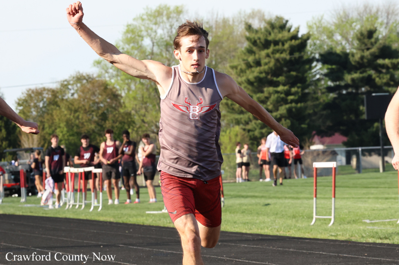 Male sprinter in a gray tank top and red shorts racing on an outdoor track, with teammates and spectators in the background.