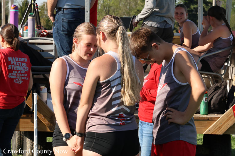 Group of young athletes in gray uniforms huddled and talking on a sunny outdoor track setup with equipment and onlookers in background.