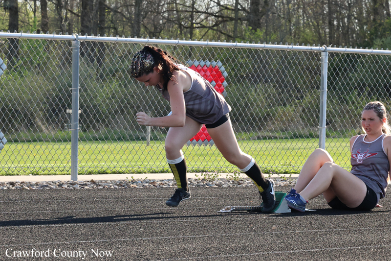 Sprinter in a gray tank top runs on an outdoor track while a teammate sits on the ground nearby by starting blocks and a chain-link fence behind them.