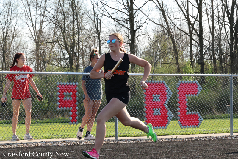 Female sprinter in a black uniform runs on an outdoor track, neon-green shoes, with a chain-link fence and bystanders behind.