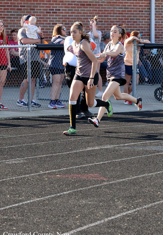 Two female runners in gray uniforms sprint on a track, with spectators behind a chain-link fence watching the race.