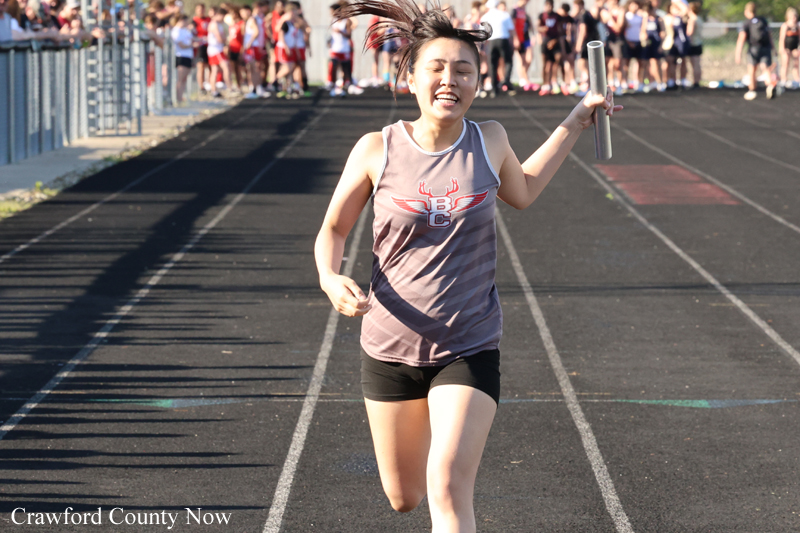 Female runner crosses the finish line on a track, baton in hand, as spectators cheer in the background.