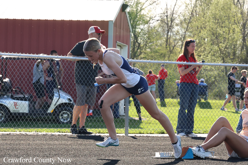 Female sprinter in white and blue starts a race from the blocks at an outdoor track meet, with onlookers behind a chain-link fence.