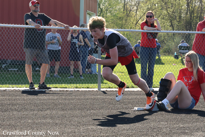 Young male athlete in mid-sprint leaping over a hurdle on an outdoor track as spectators watch behind a chain-link fence.