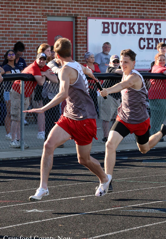 Two male relay runners sprint on a track as one passes a baton, with spectators watching behind a chain-link fence near a Buckeye Center sign.