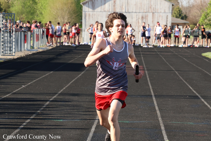 Relaying runner in a gray tank top and red shorts sprinting on an outdoor track with a baton, as spectators watch in the background.