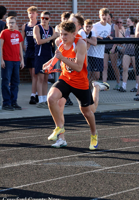 Relay race moment: athlete in orange jersey sprinting on track with baton while spectators watch behind a chain-link fence