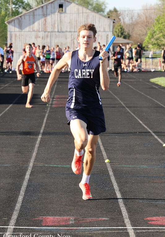 Male relay runner in a navy Carey uniform sprints on a track with a blue baton, spectators and a barn in the background.