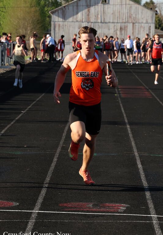 Male relay runner in an orange Seneca East uniform sprints on a track holding a baton, with spectators and a barn in the background.
