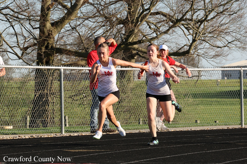 Two young female runners in white uniforms sprint side by side on an outdoor track, with spectators behind a chain-link fence.