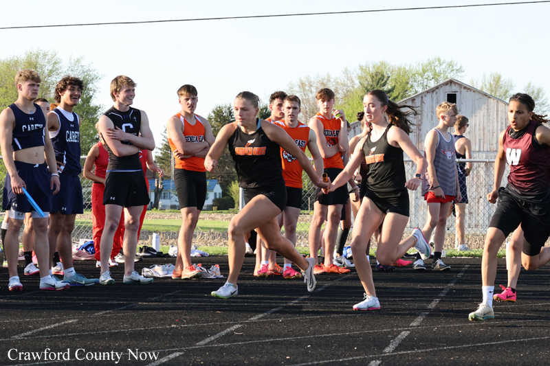 Group of high school athletes sprinting from the starting line on an outdoor track, with teammates watching in the background.