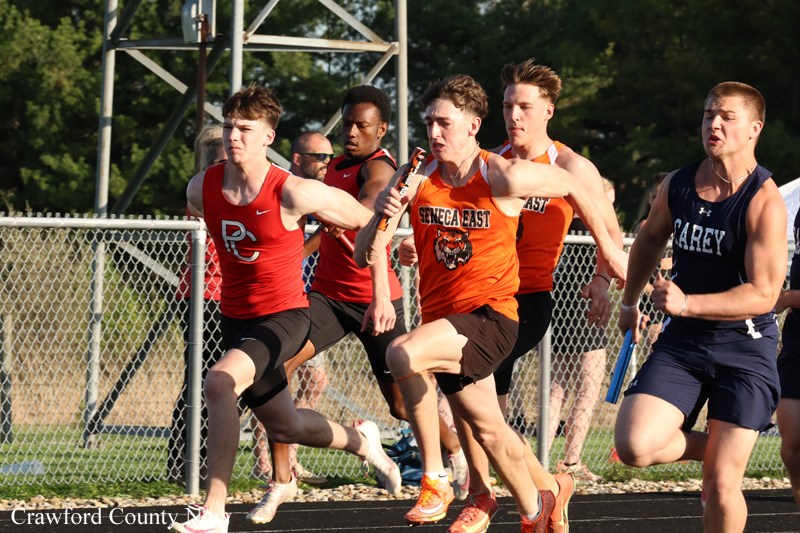 Runners in a relay race sprint on a track; the lead athlete in orange passes the baton to a teammate amid other sprinters behind him.