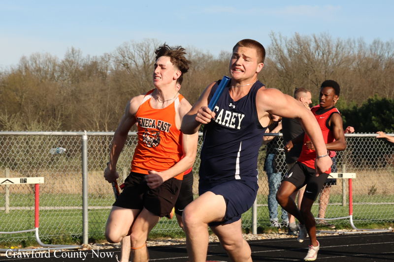 Two male sprinters in a relay on an outdoor track; the front runner in navy holds a blue baton, while an orange-uniform racer stays close behind.
