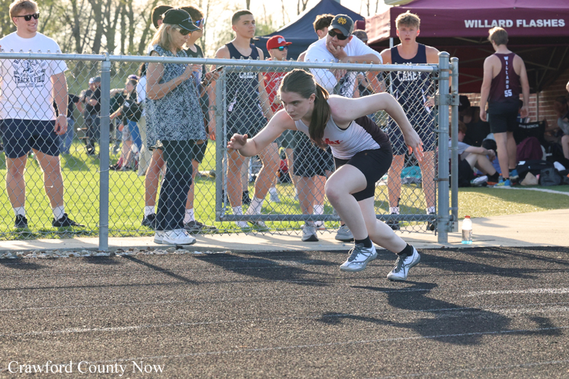 Female sprinter in white top and black shorts crouches at the start on an outdoor track with spectators behind a fence.