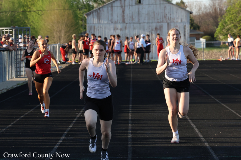 Three female sprinters race on an outdoor track with a small crowd in the background; two wear white shirts with red logos.