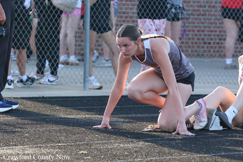 Female sprinter crouched at the starting line on a track, ready to race, with spectators behind a chain-link fence.