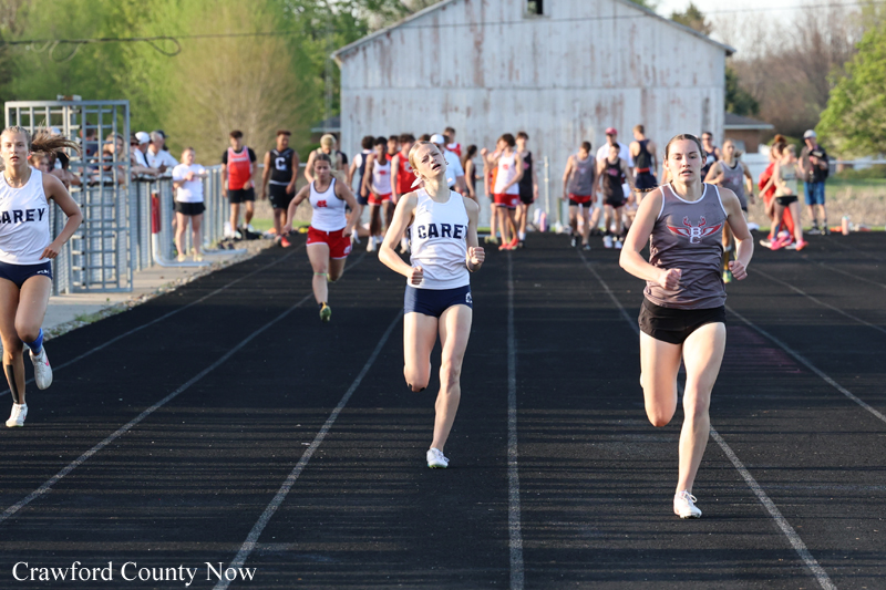 Girls' track race on an outdoor track; three runners lead with a crowd along the fence and a weathered barn in the background. Watermark 'Crawford County Now' bottom left.