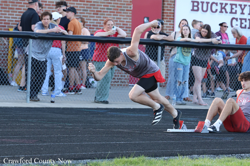 Male sprinter in a gray tank top and red shorts launches from starting blocks on a track as spectators watch from behind a chain-link fence.