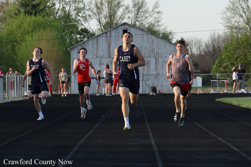Four male sprinters race down a track toward the finish, with a small crowd and a weathered barn in the background.