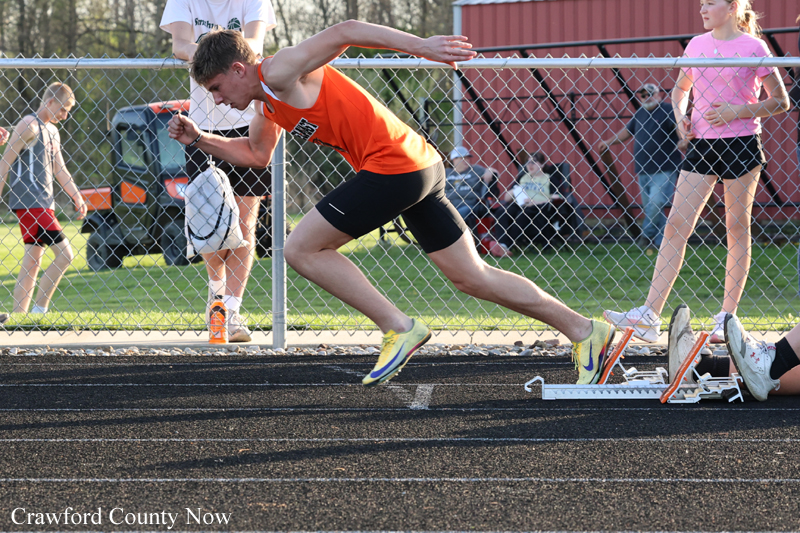 Male sprinter in orange top and black shorts leaps from starting blocks on a track, with spectators behind a chain-link fence.
