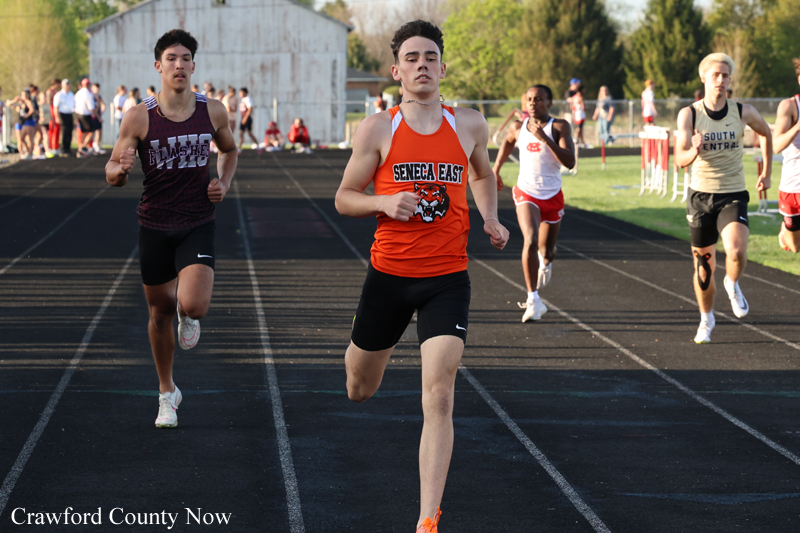 Young male sprinter in orange Seneca East uniform leads a race on an outdoor track, with competitors close behind and spectators in the background.