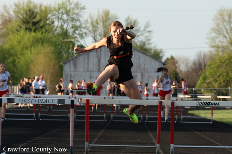 Female sprinter in a black uniform jumps over a hurdle on an outdoor track, with spectators and a barn in the background.