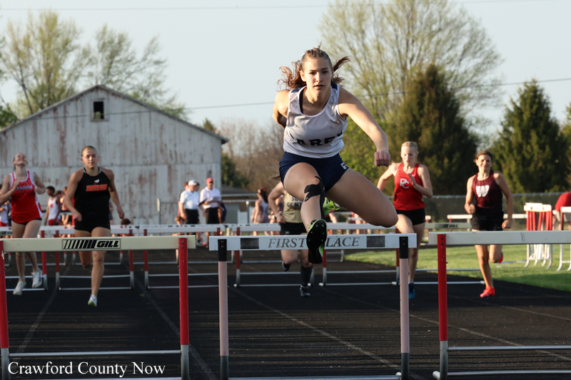 A female hurdler clears a hurdle mid-jump on an outdoor track, others running behind him, with a barn and trees in the background.