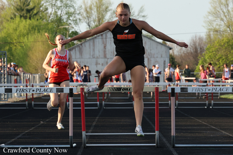 Female hurdler in a black uniform clears a hurdle mid-race on a track, with another runner in red behind and spectators in the background.