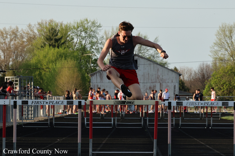 Male sprinter mid-air clearing a hurdle on an outdoor track, with spectators in the background.
